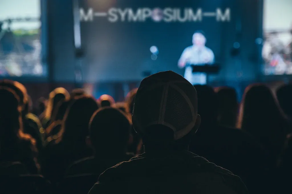 Audience watching a speaker on stage at a darkly lit symposium or conference, with the word “SYMPOSIUM” visible on a blurred screen in the background.